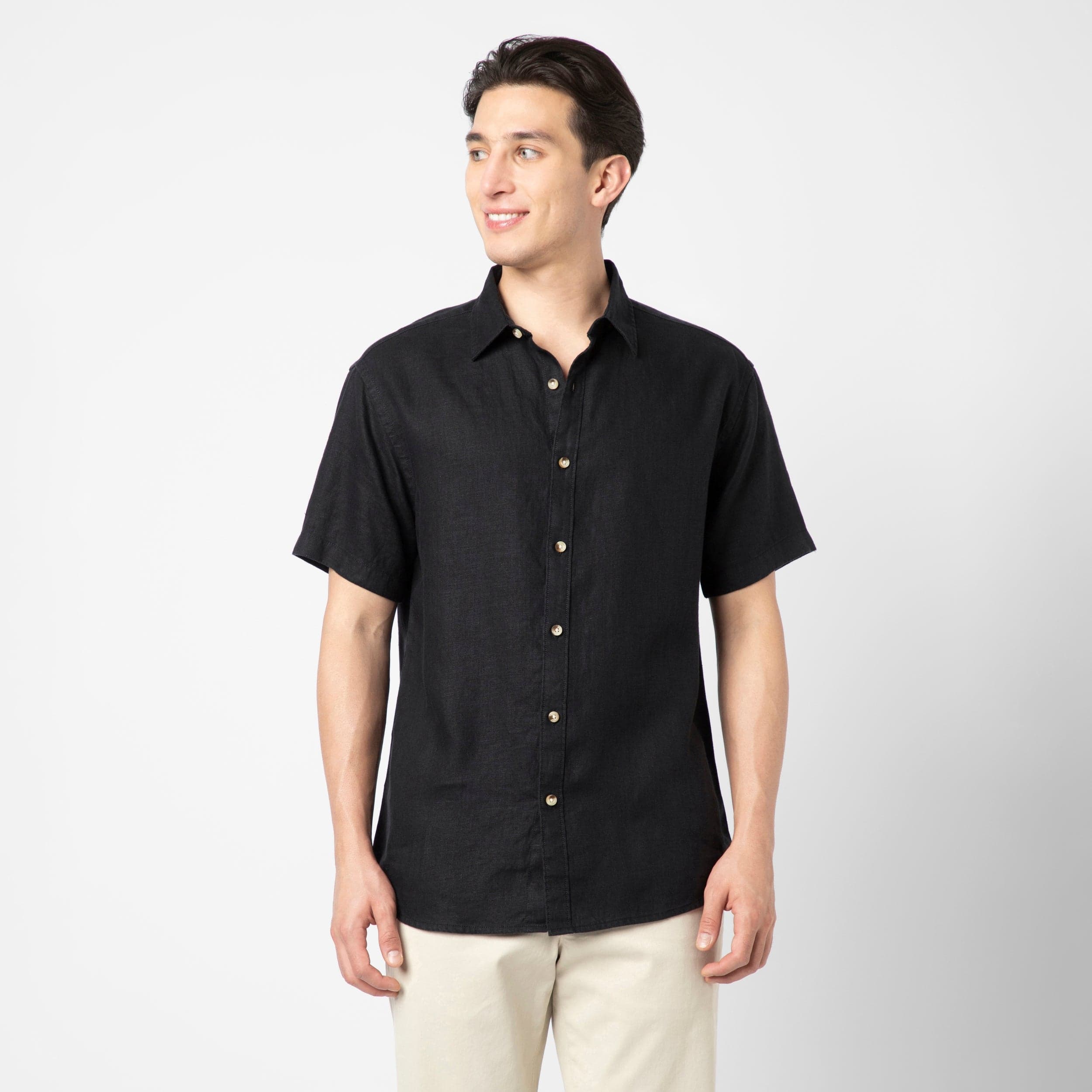 A man smiles and looks to the side against a plain white background, wearing the Linen Short Sleeve Shirt in Black with light pants, highlighting natural comfort and effortless style.