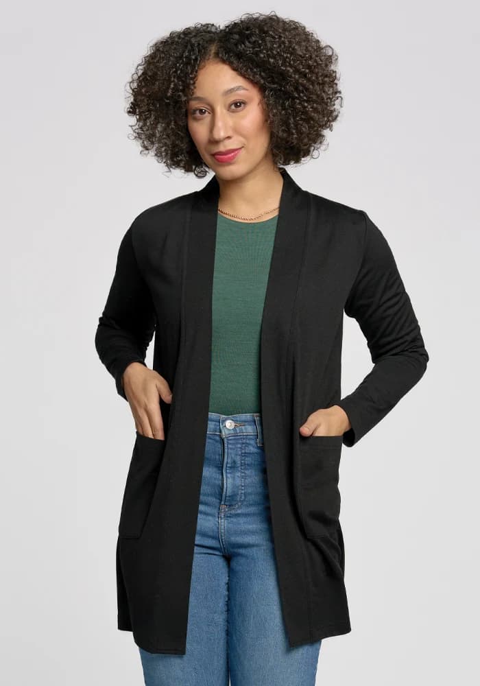 A curly-haired person wears a Woolx Paisley Cardigan in black over a green top and blue jeans. Made from lightweight Merino wool, they stand with hands in the cardigan pockets against a plain background. | Tori is 5'7", wearing a size S