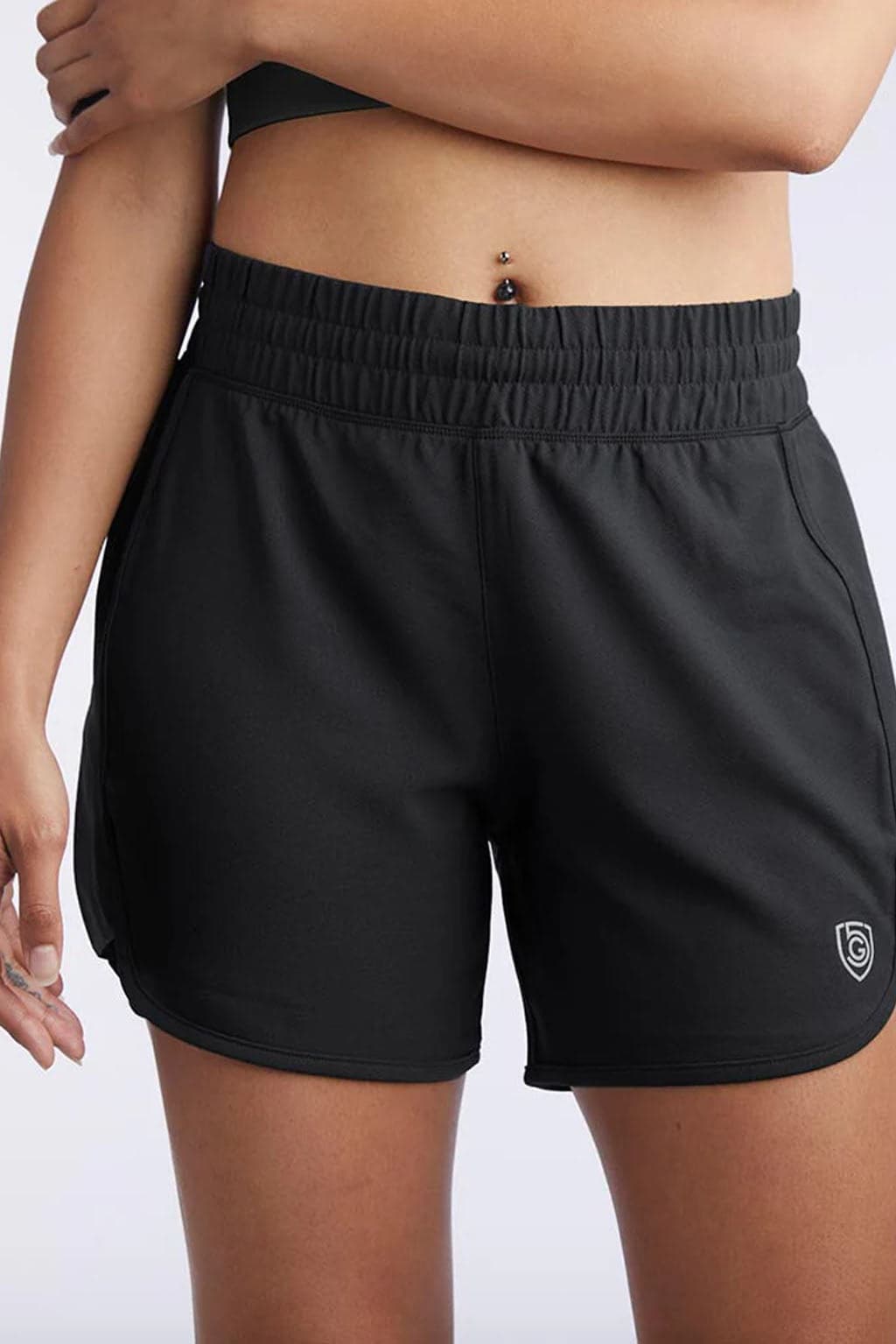 A person wears black Indie Women's Sport Shorts featuring an elastic waistband and a small logo on the left thigh, paired with a black crop top. Four-way stretch fabric ensures comfort for any activity; a navel piercing is visible.