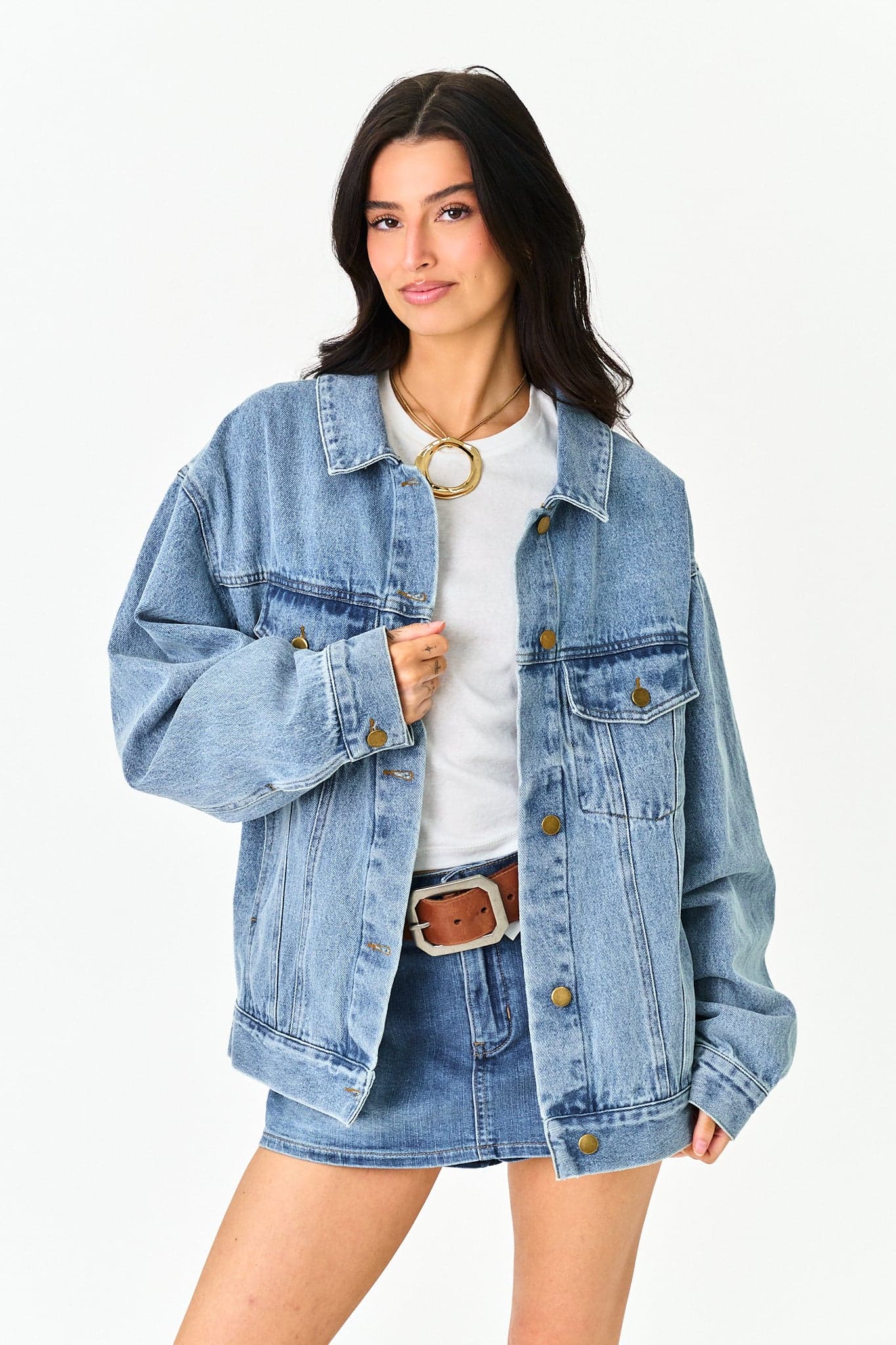A woman modeling an oversized light-wash blue denim jacket over a white top and denim skirt, with a brown belt and gold ring necklace, against a plain white studio background.
