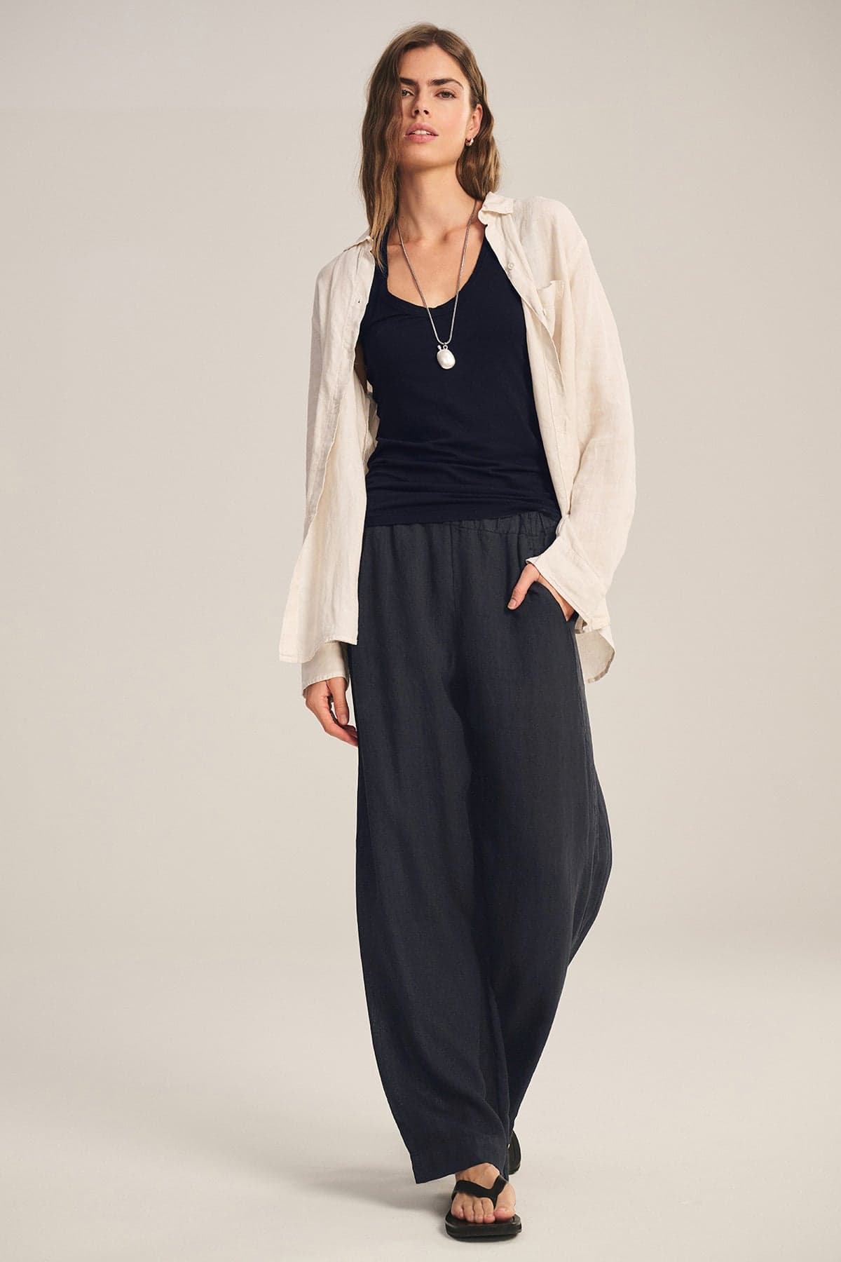 A person stands in a neutral studio wearing the LOLA LINEN PANT by Velvet by Graham & Spencer, styled with a cream shirt over a black top, black sandals, and a necklace.